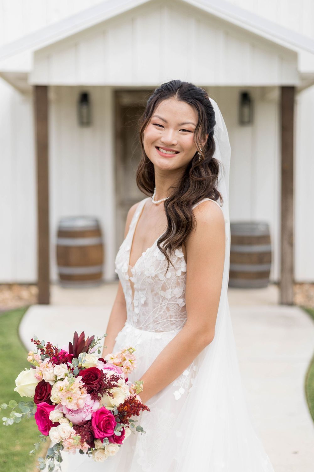 Smiling bride in lace wedding gown holding burgundy and pink bouquet, wearing veil, standing outside white barn venue in Chappell Hill, TX
