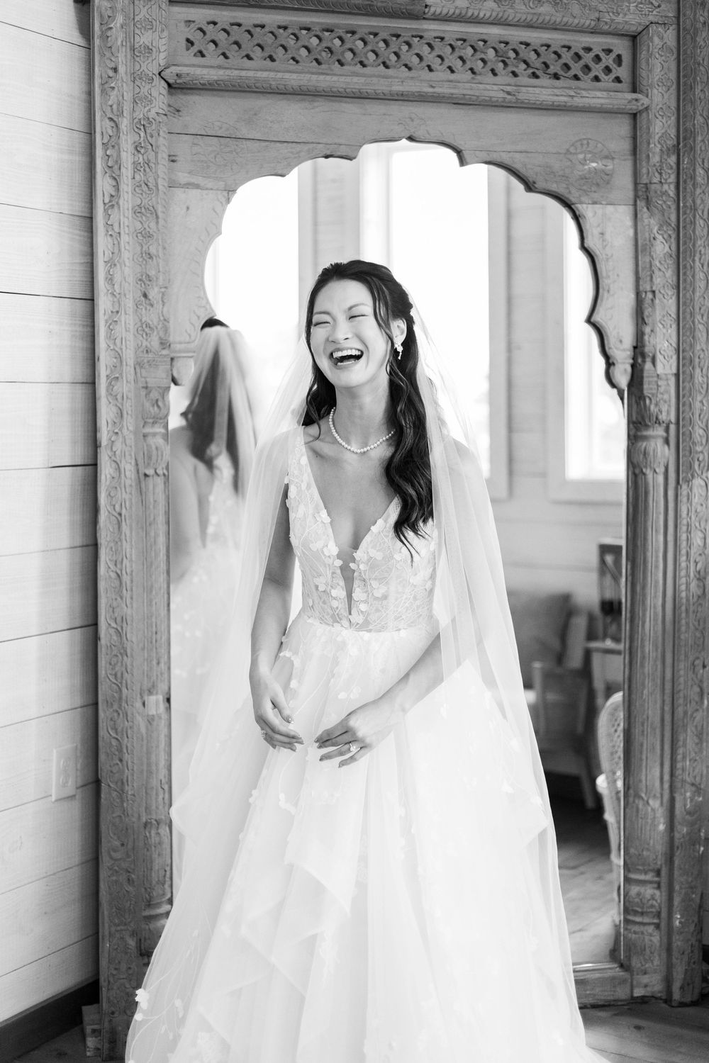 Joyful bride in lace wedding gown and veil laughing in black and white photo framed by ornate mirror.