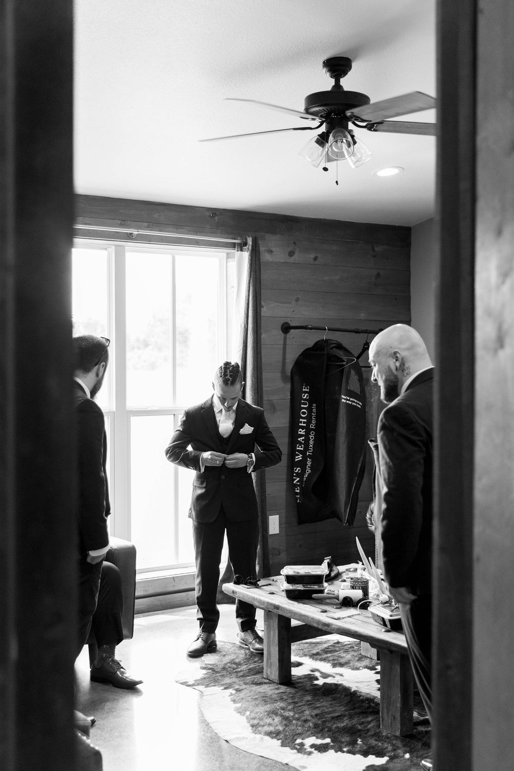 Black and white photo of groomsmen helping groom button jacket before wedding ceremony in rustic room.