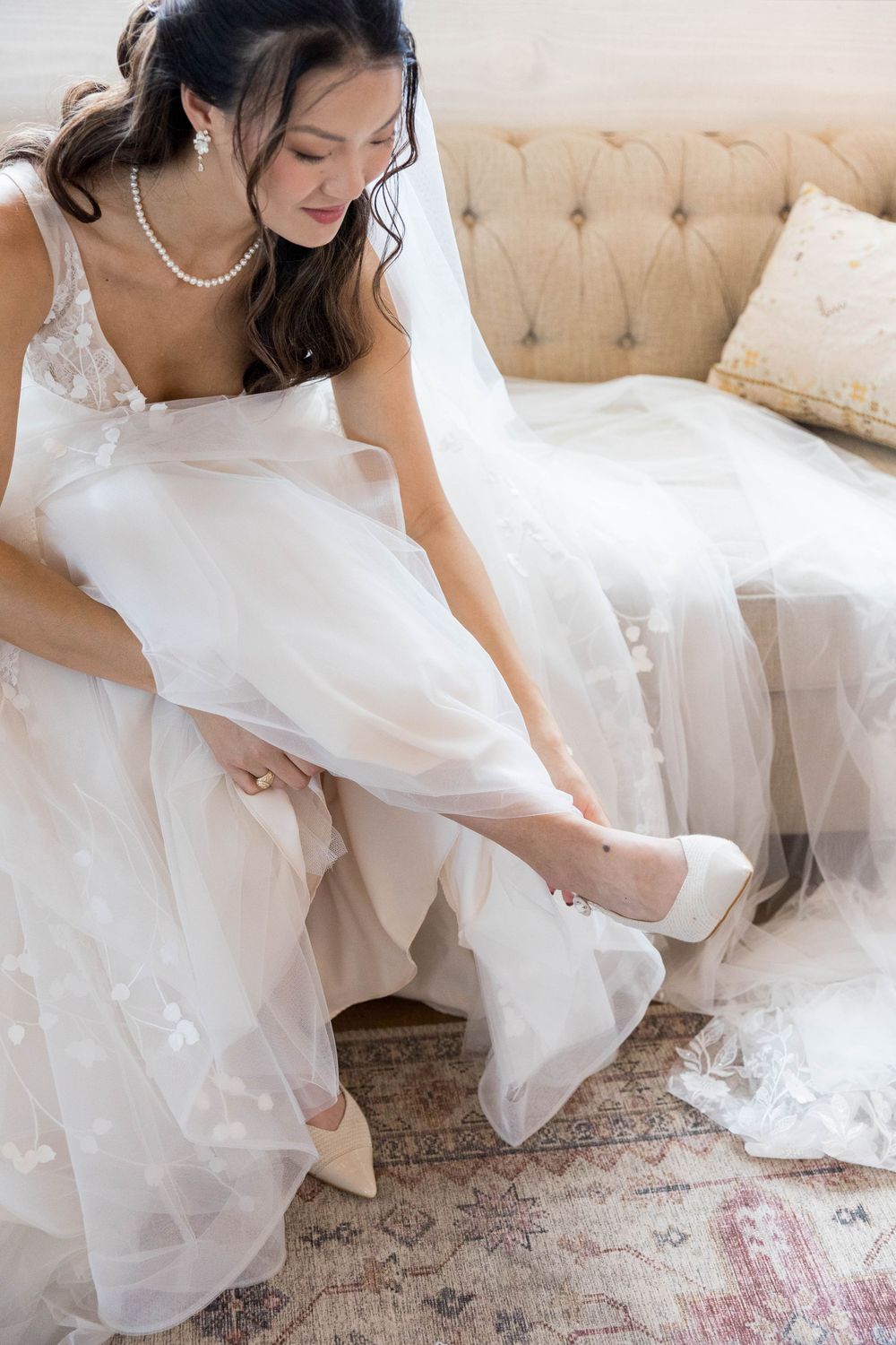 Bride in white lace gown and veil putting on heels while seated on a tufted beige sofa.