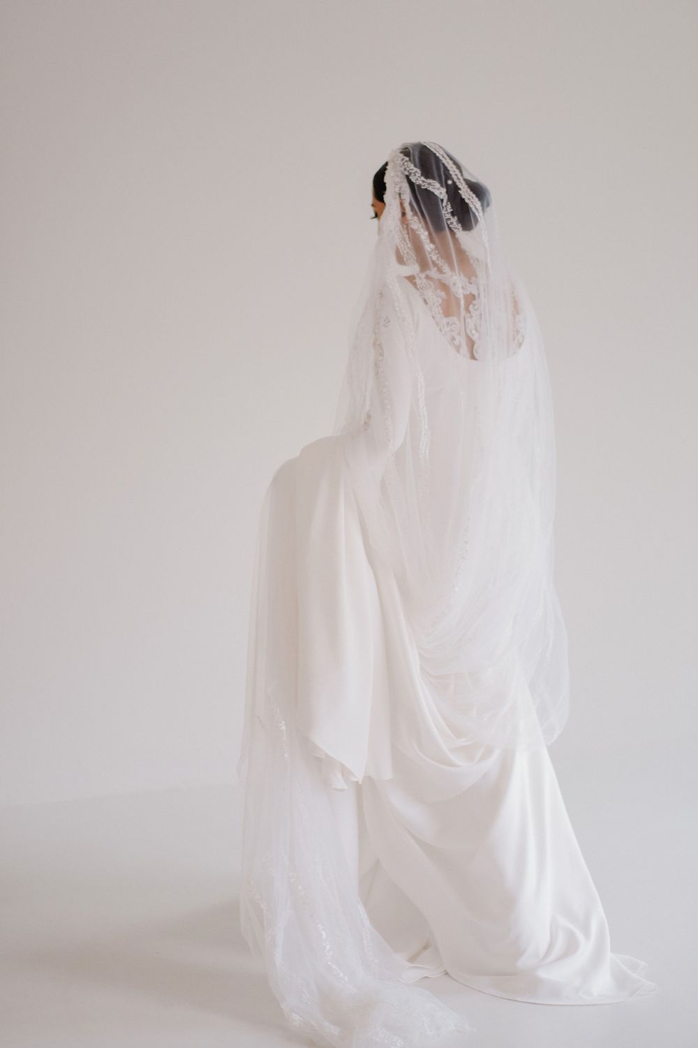 Bride seen from behind wearing flowing white wedding gown and long delicate veil against a clean white background.
