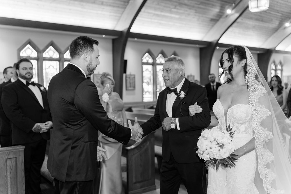 Groom shaking hands with father of the bride during black and white church wedding ceremony.