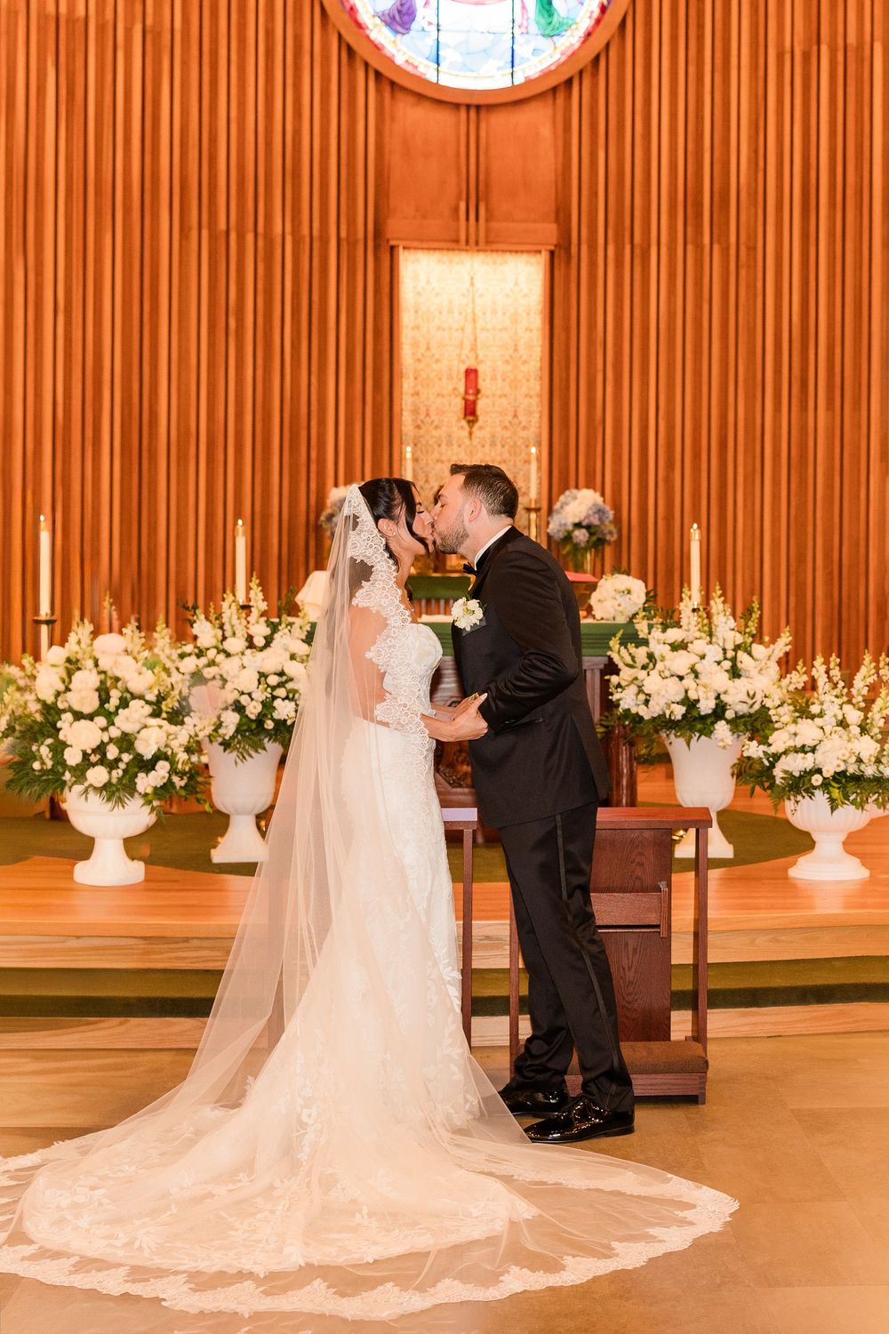 Bride and groom share first kiss at church altar surrounded by white floral arrangements and candles.