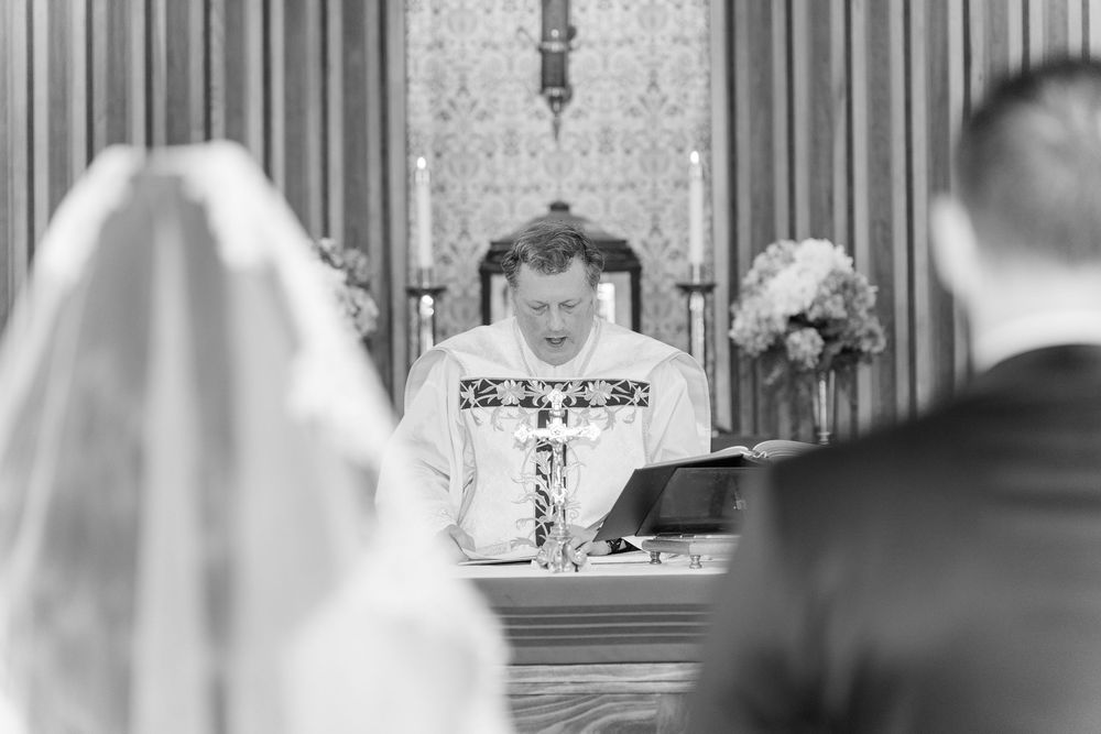 Priest reads at altar during wedding ceremony in church with bride and groom seated before him.