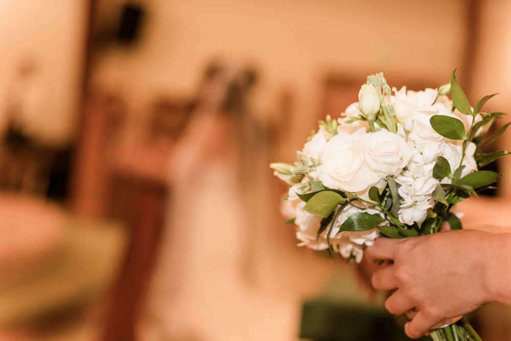 Groom holds white rose and greenery bridal bouquet inside wedding venue with bride visible in background.