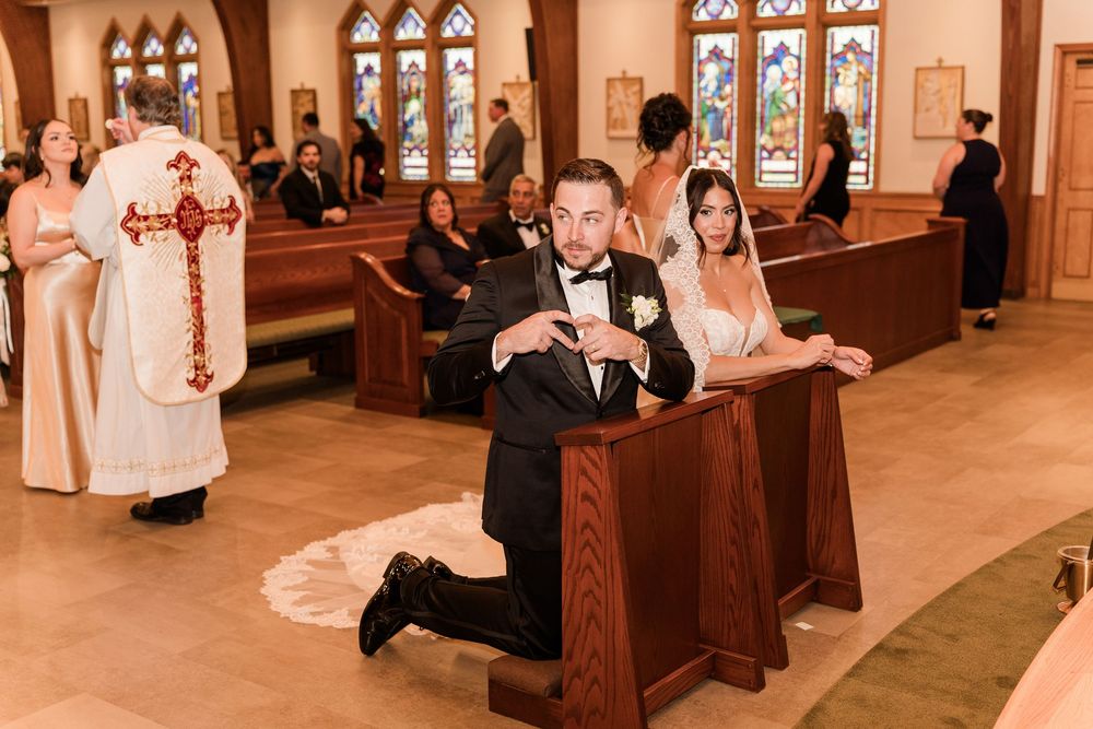 Bride and groom kneel at church pew during wedding ceremony with guests and priest in background.