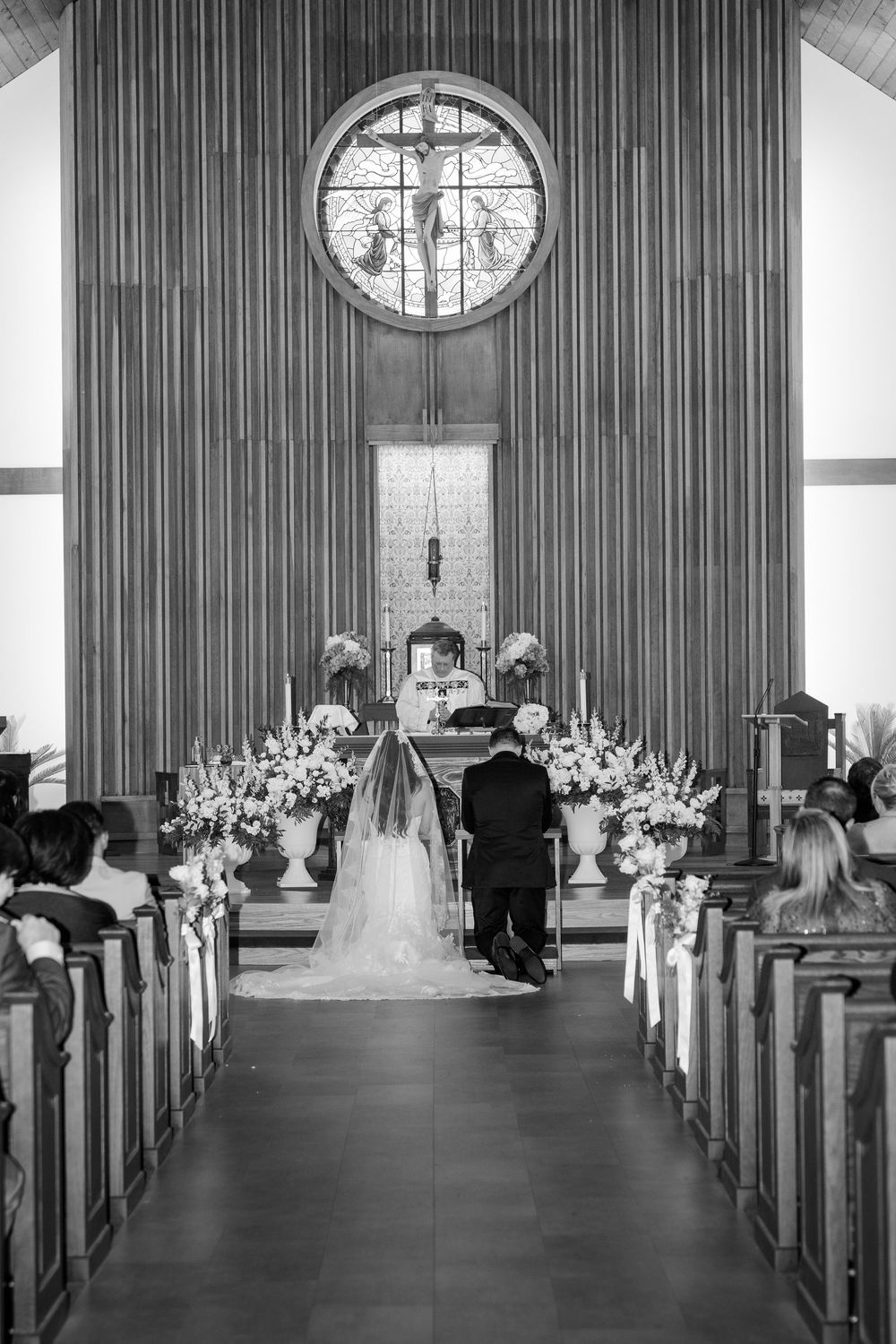 Bride and groom kneeling at altar in elegant church wedding ceremony with floral arrangements.