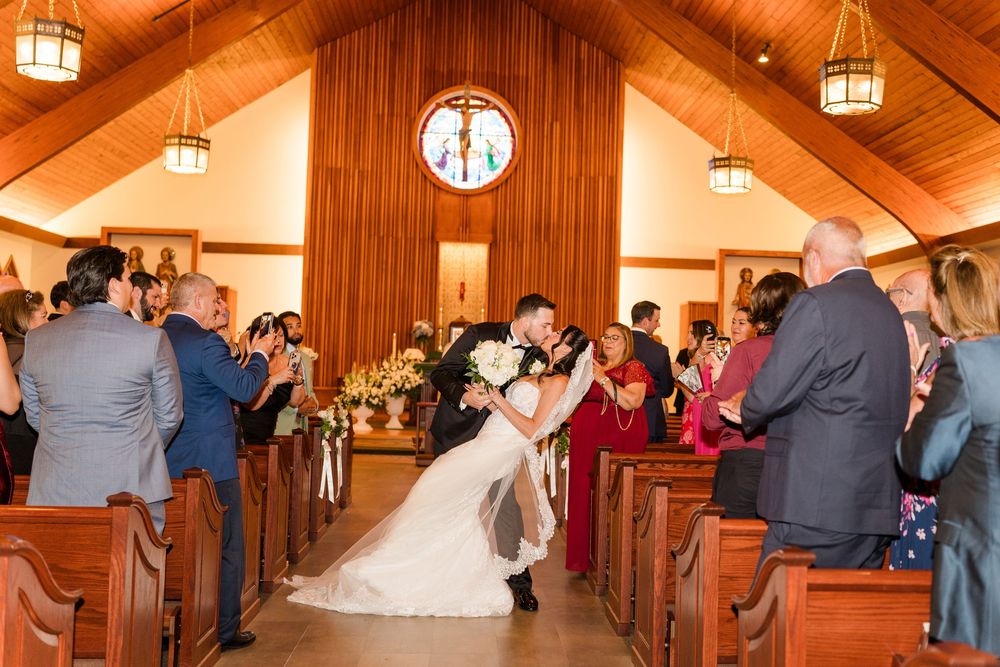 Newlyweds sharing romantic kiss in church aisle surrounded by cheering wedding guests.