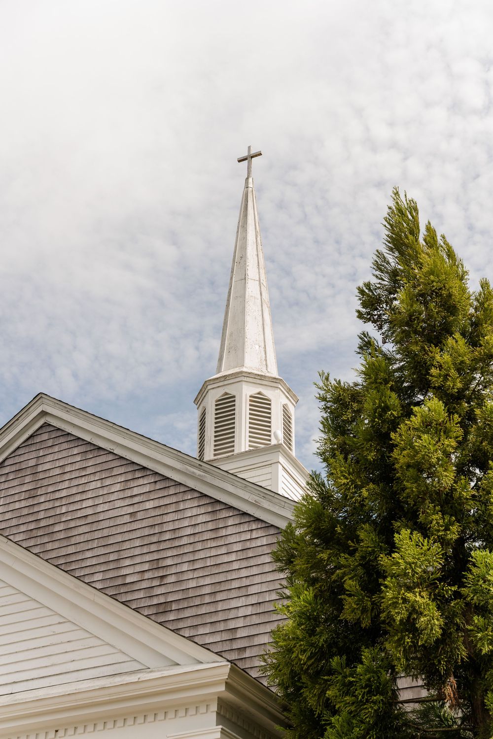 White wooden church steeple with cross atop a shingled roof, framed by a green evergreen tree against a cloudy sky.