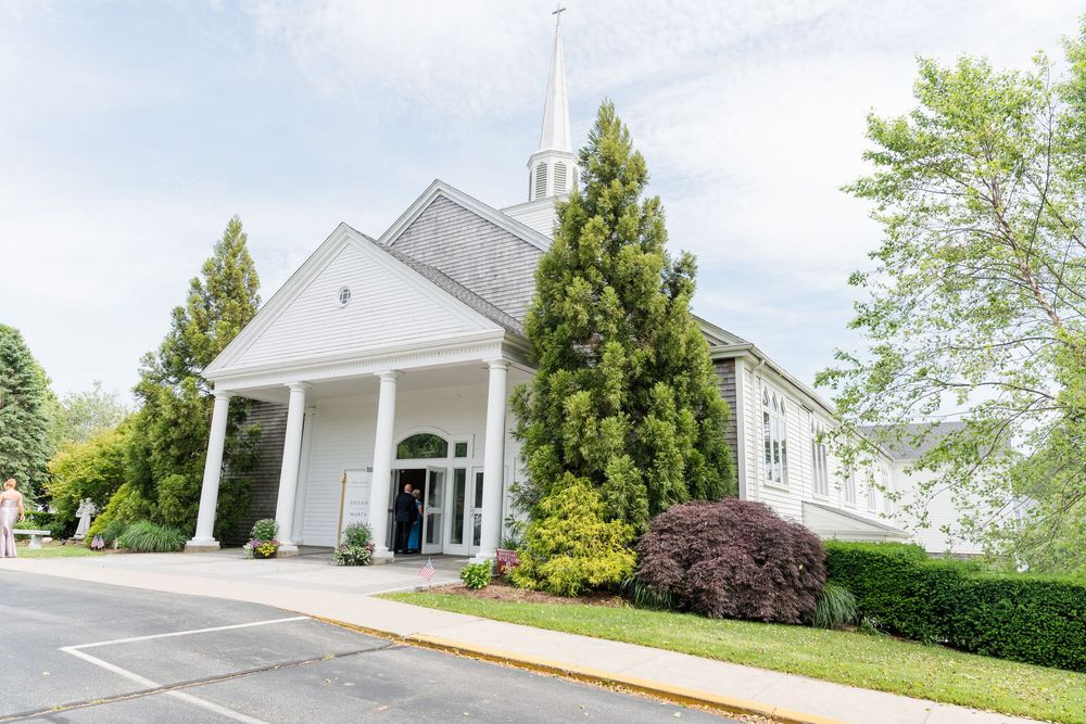 White church with steeple, columned entrance, and lush green trees on a sunny day.