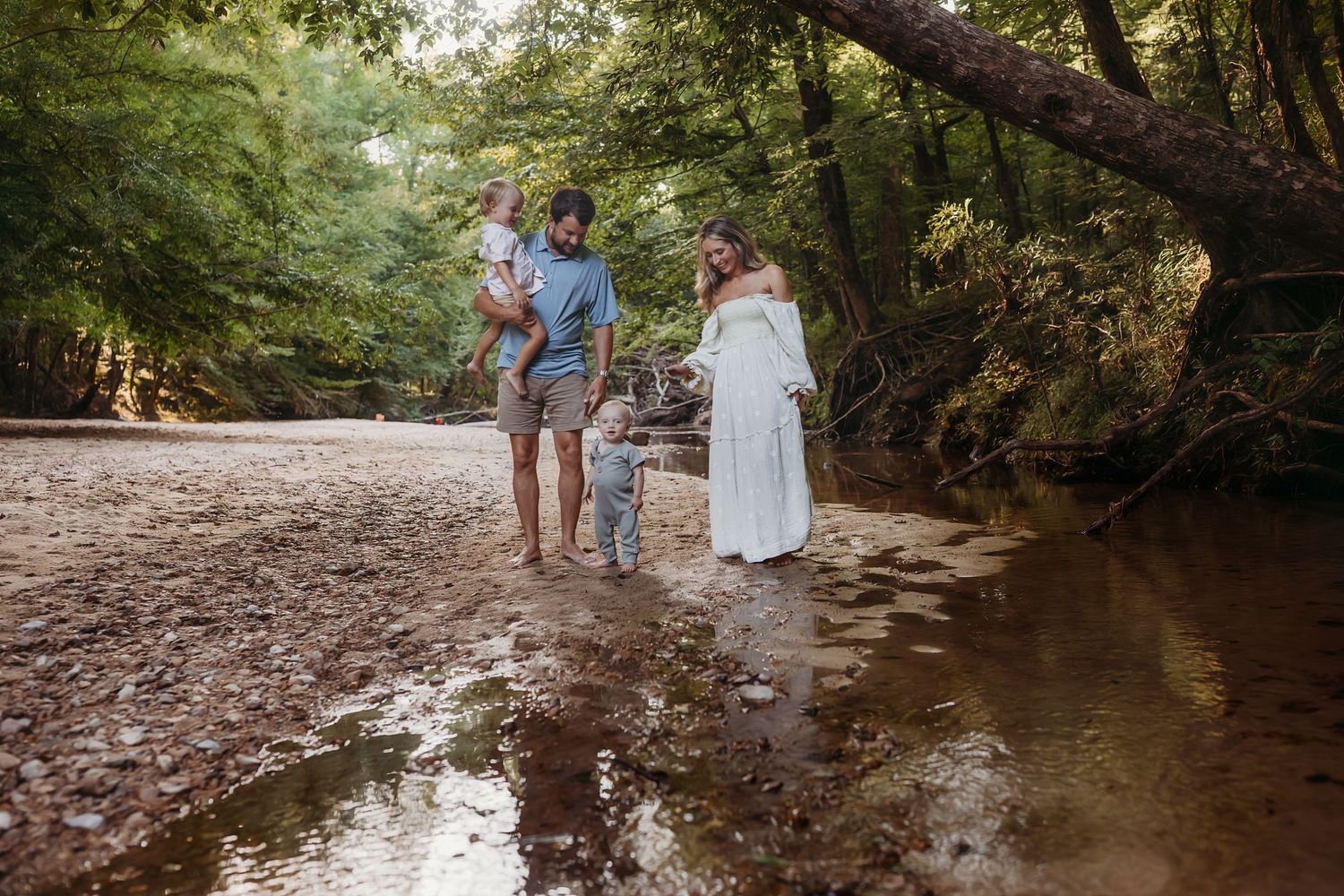 Family of four enjoying a peaceful walk along a forest creek, with parents and two young children surrounded by lush green trees.