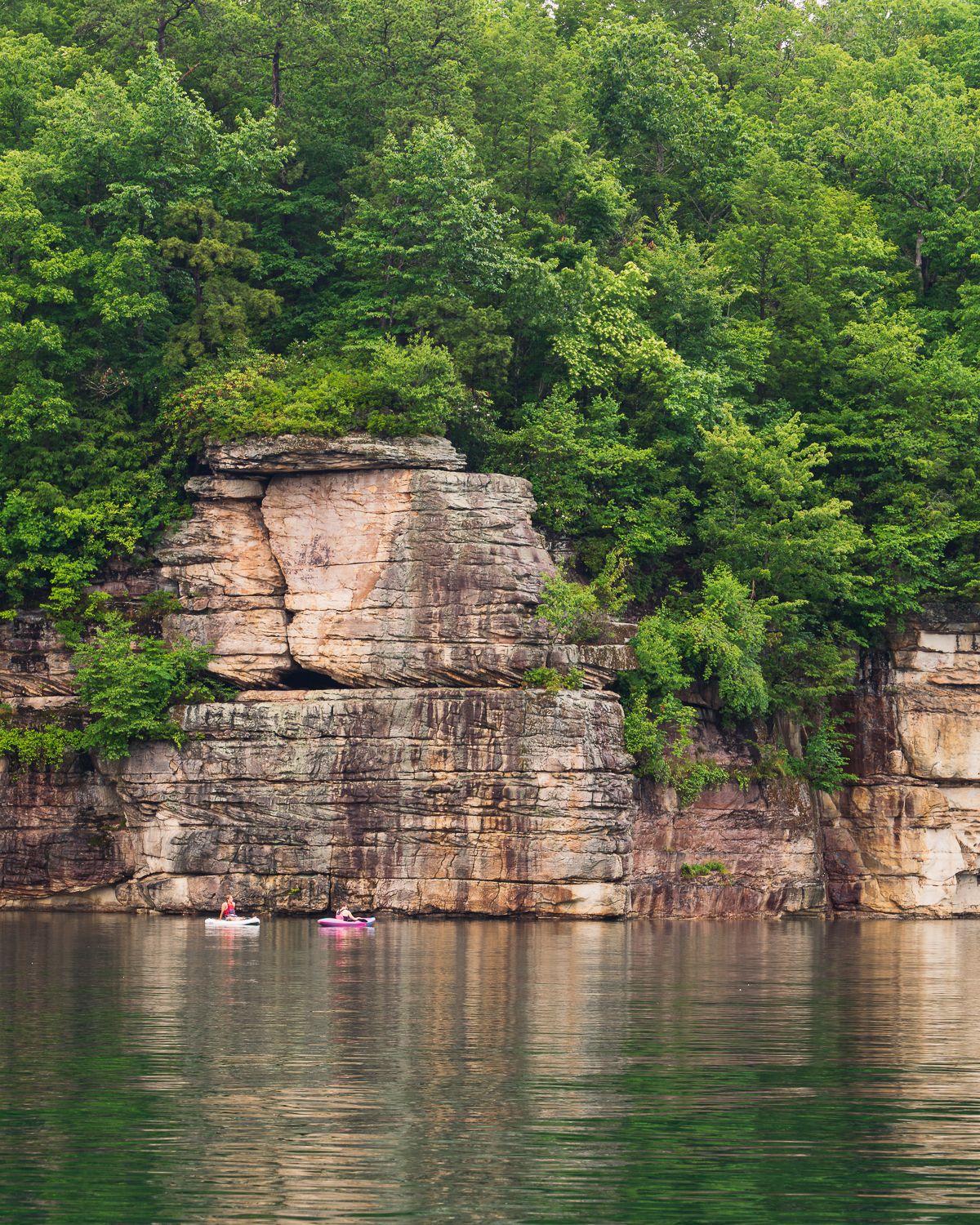 Kayakers paddle on emerald green water beside towering limestone cliffs covered with lush green trees.