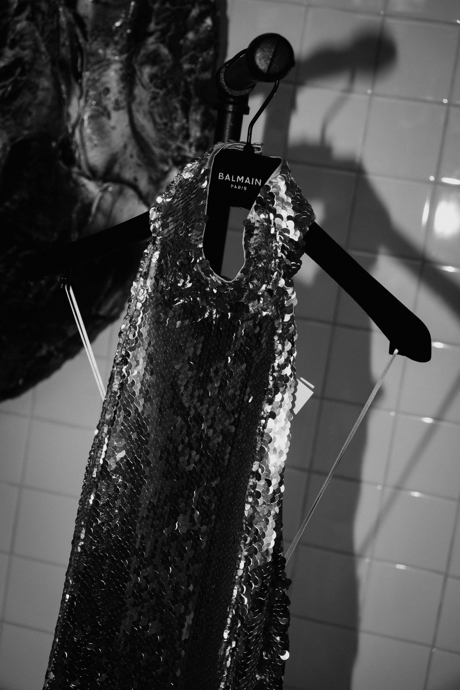 Black and white photo of a sequined Balmain Paris dress on a velvet hanger against a tiled wall.