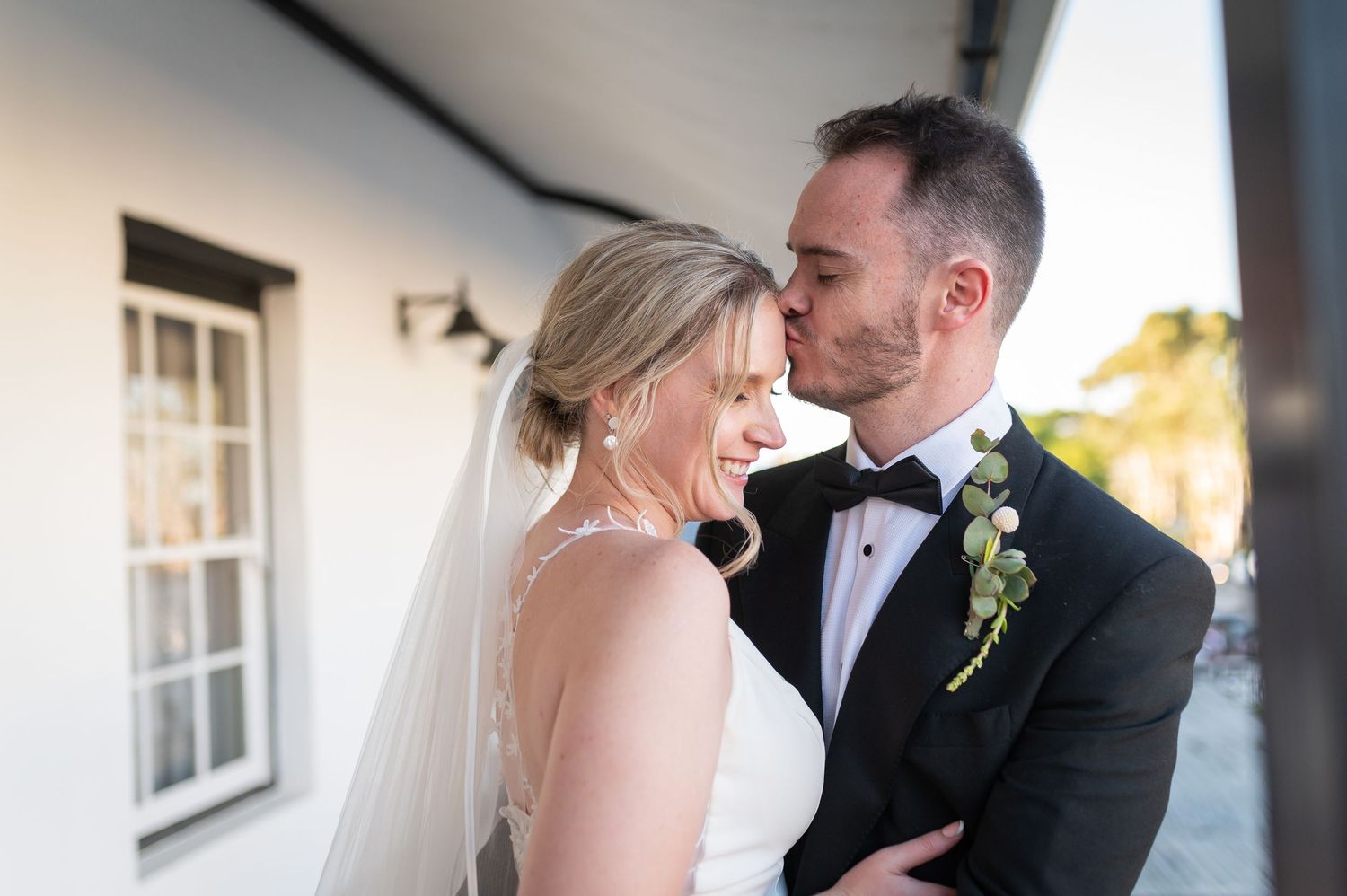 Bride and groom share a romantic moment outdoors, groom in tuxedo kissing bride's temple as she smiles in white gown.