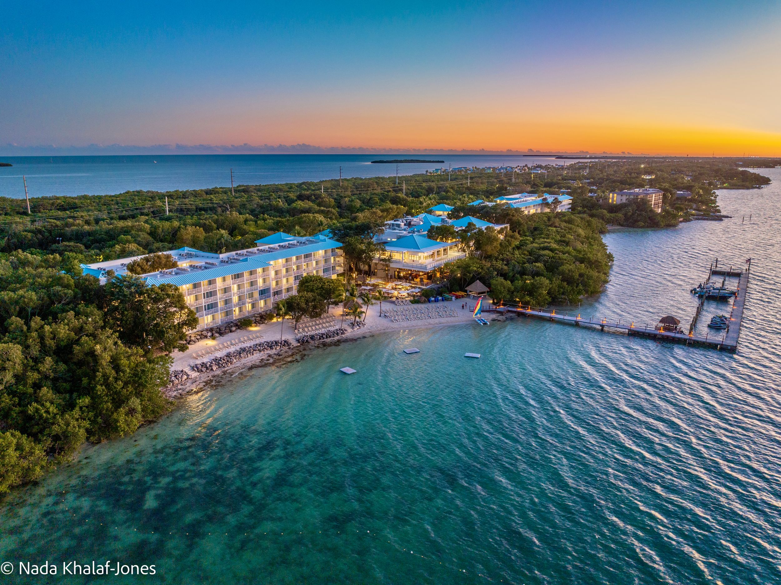 Aerial sunset view of a tropical island resort with turquoise waters, lush trees, and a private dock.