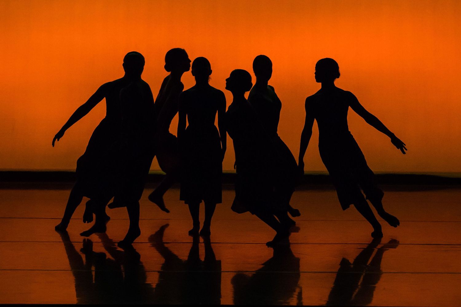 Silhouettes of five dancers performing gracefully against a vibrant orange background on a reflective stage.