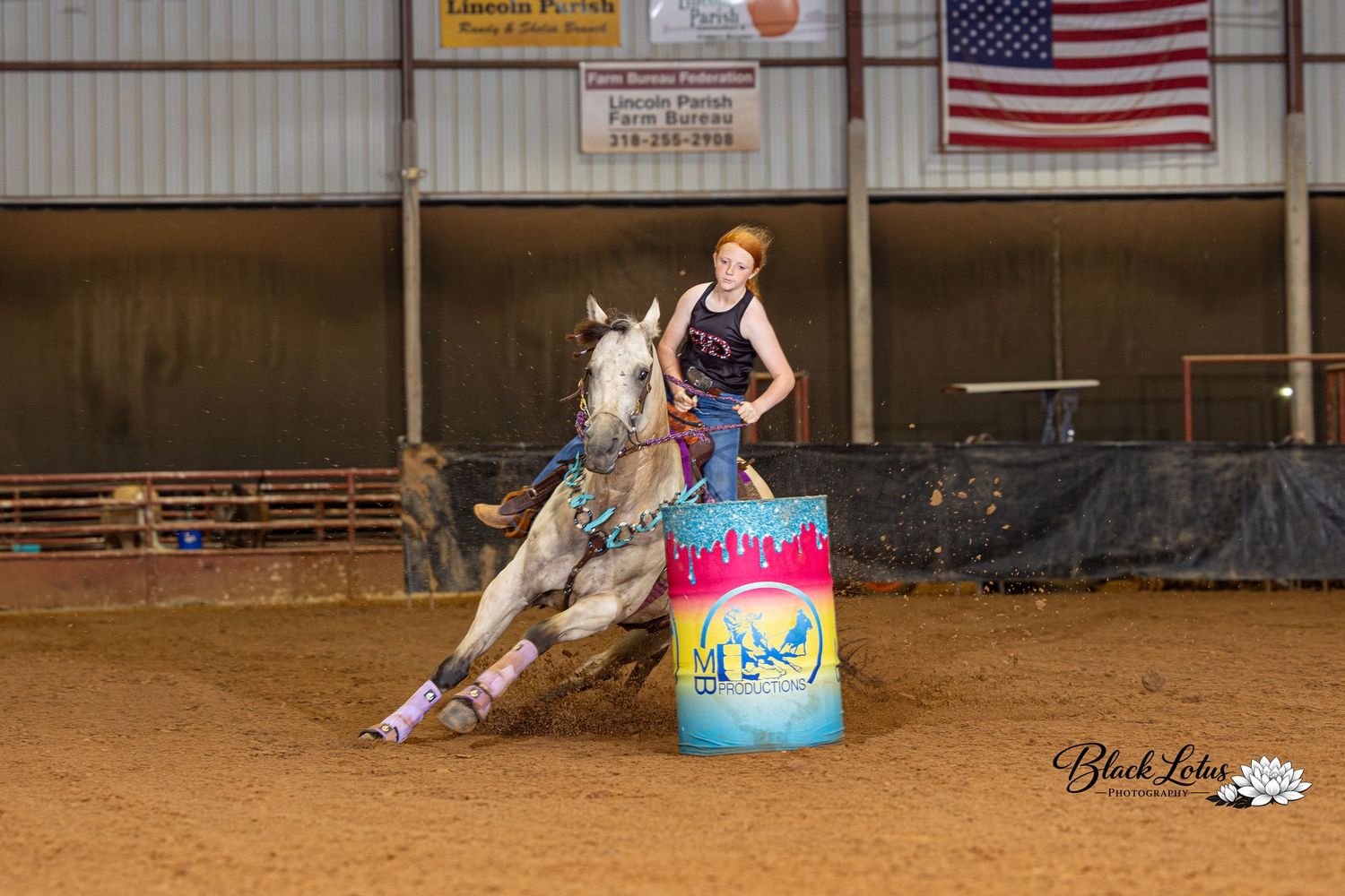 Young girl competing in barrel racing on a gray horse at an indoor rodeo arena.