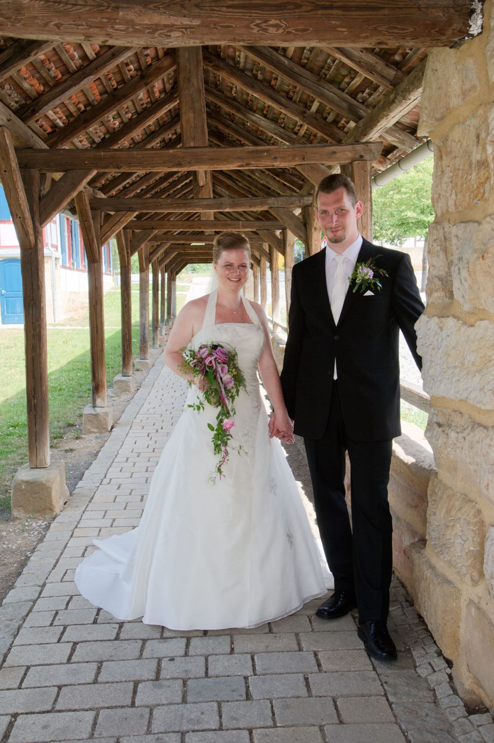 Brautpaar spaziert Hand in Hand durch einen überdachten historischen Gang. Klassische Hochzeitsfotografie am Standort Eggolsheim.