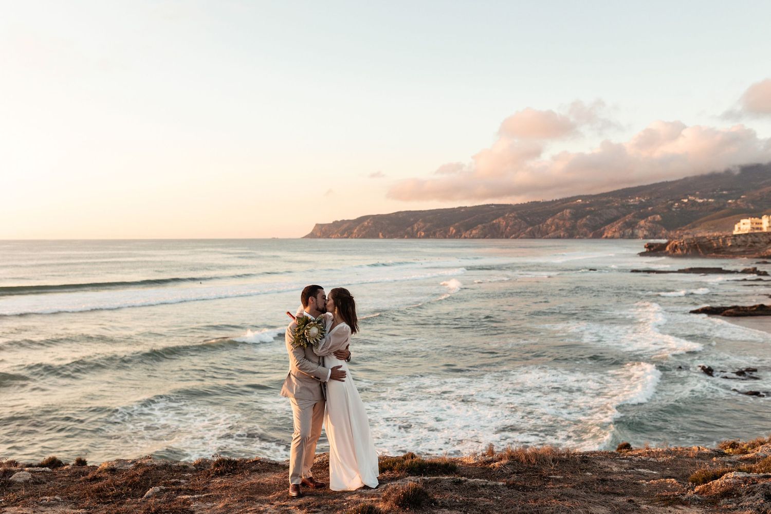A couple embraces on a rocky coastline at sunset with waves crashing against the shore and mountains in the background.