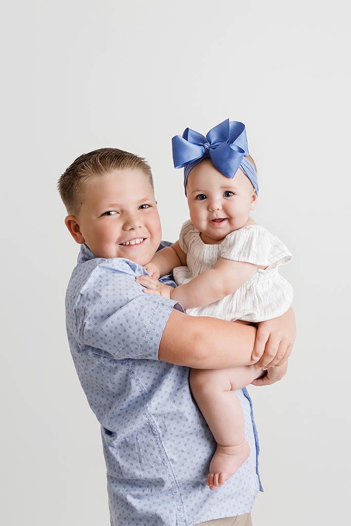 A smiling boy holds a baby girl wearing a blue bow headband, both dressed in blue and white outfits against a white background.
