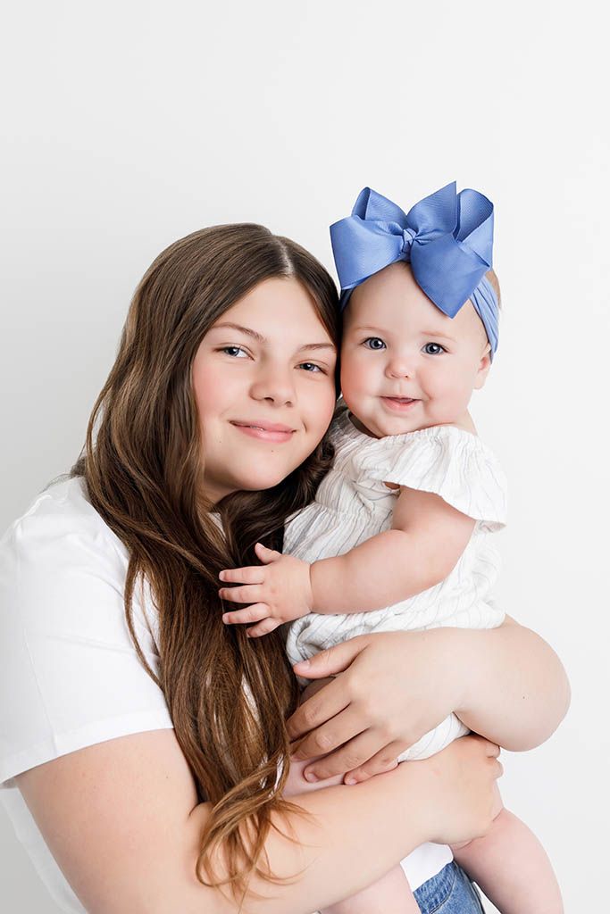 Teen girl with long brown hair holds smiling baby wearing a blue bow headband, posing together against white background.