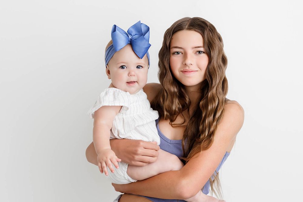 A teenage girl holds a smiling baby wearing a blue bow headband against a white background.