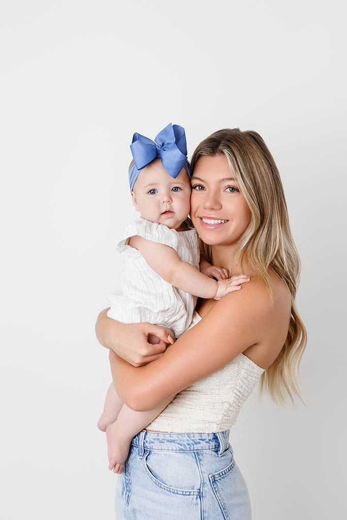 Smiling young woman holding a baby girl wearing a blue bow headband, posing together against a white background.