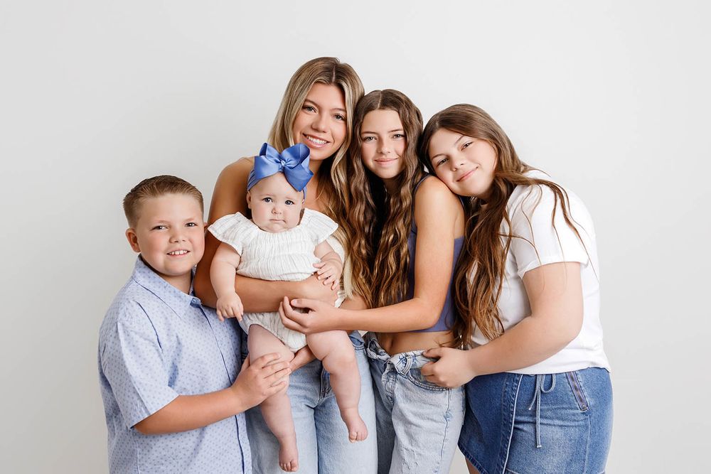 A smiling mother poses with her four children, including a baby in a blue bow, against a white background.
