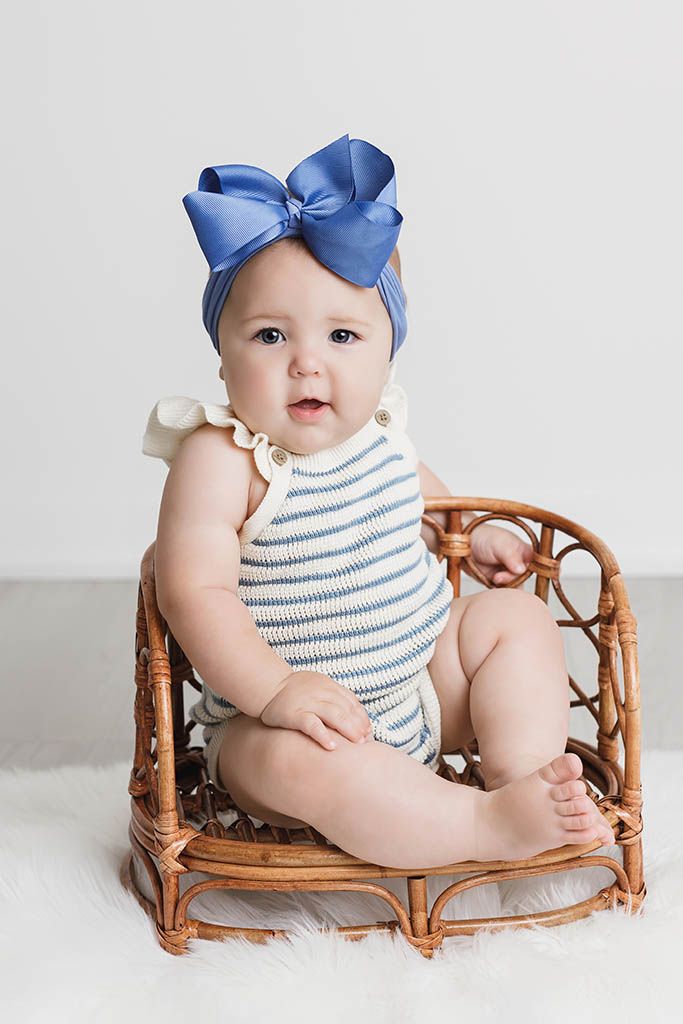 Baby girl in striped romper and blue bow headband sitting in a rattan chair on a white fluffy rug.