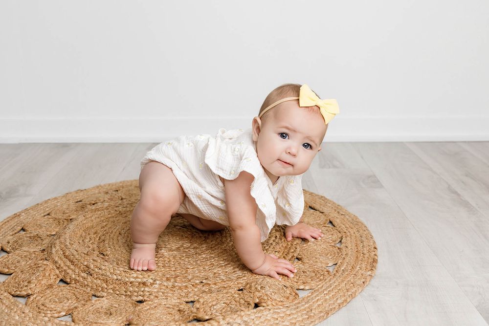 An adorable baby girl in a white romper crawls on a woven jute rug with a yellow bow headband.