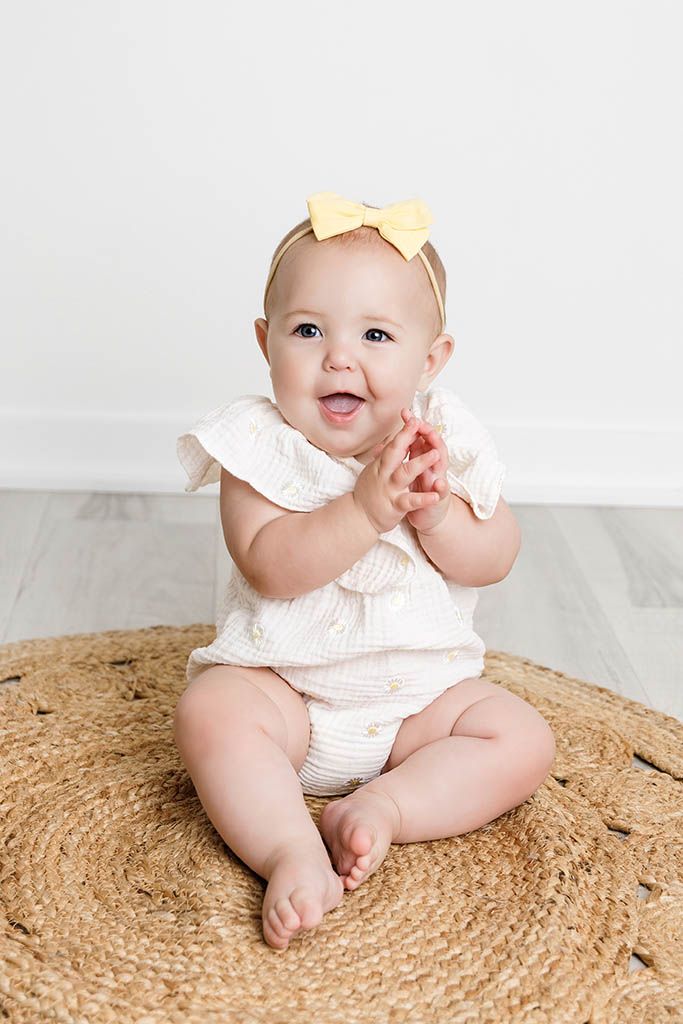 Smiling baby girl in white ruffle romper and yellow bow headband sitting on a round jute rug.