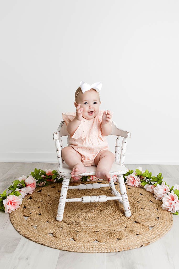 An adorable baby in a pink romper sits in a white rocking chair surrounded by pink flowers on a jute rug.