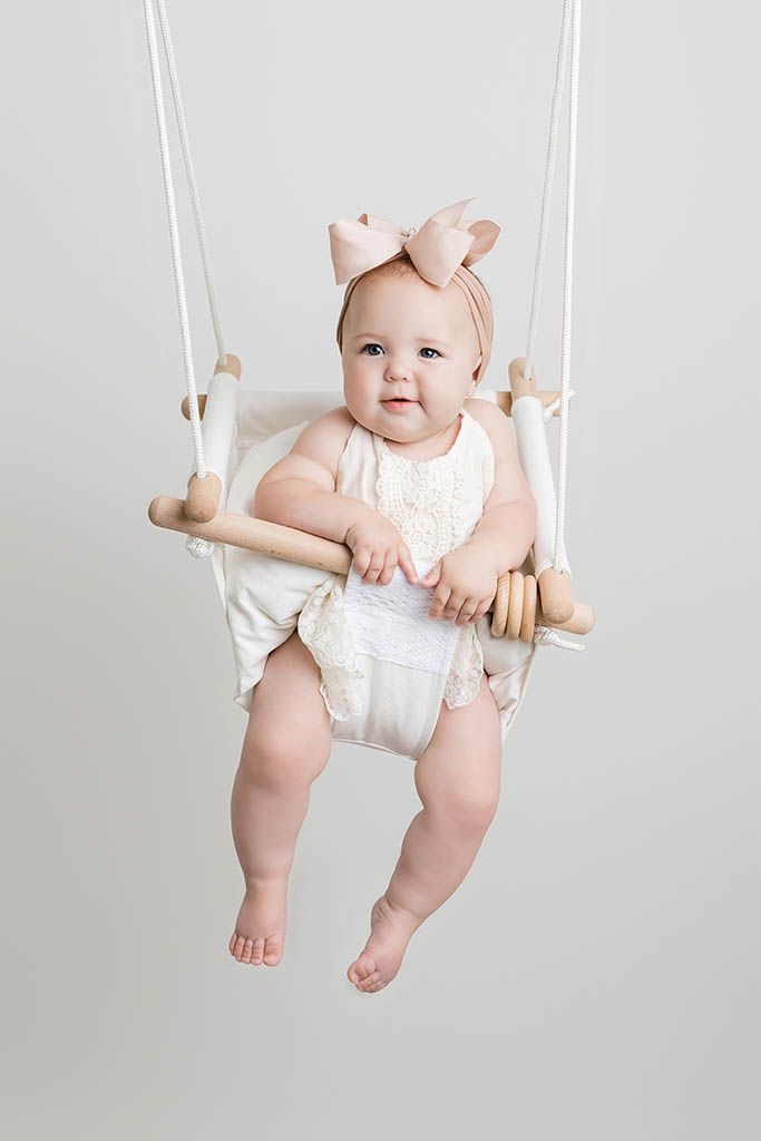 A cute baby girl sits in a hanging wooden swing wearing a cream romper and pink bow headband.