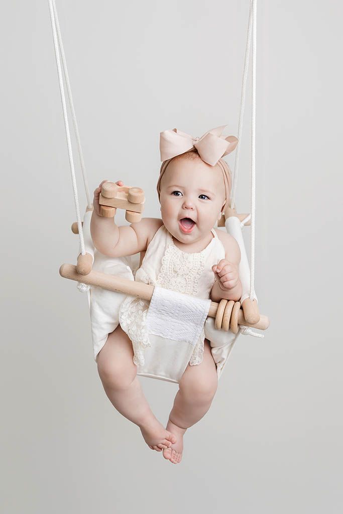 Happy baby in white lace outfit sits in a wooden swing wearing a pink bow headband.
