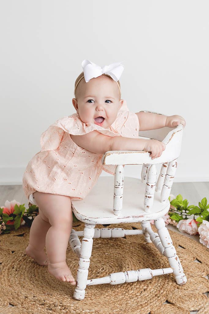 Adorable baby in peach dress with white bow stands beside a vintage white wooden chair.