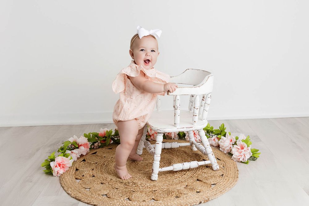 Happy baby girl in pink romper with white bow headband standing at a vintage white high chair on jute rug.