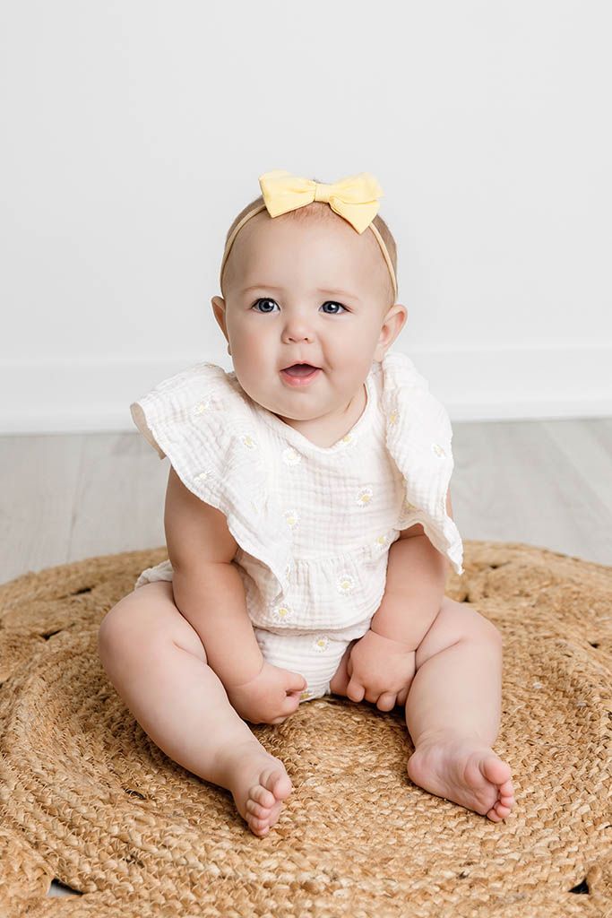 Cute baby girl sitting on woven rug wearing white ruffle romper and yellow bow headband.