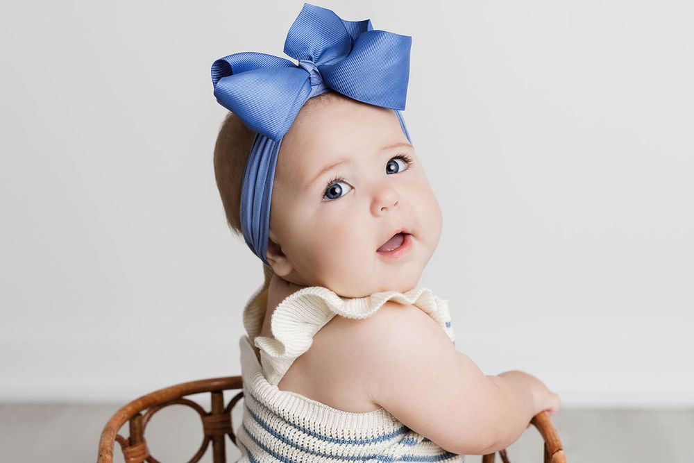 A sweet baby girl wearing a blue bow headband sits in a wicker chair in a studio portrait.