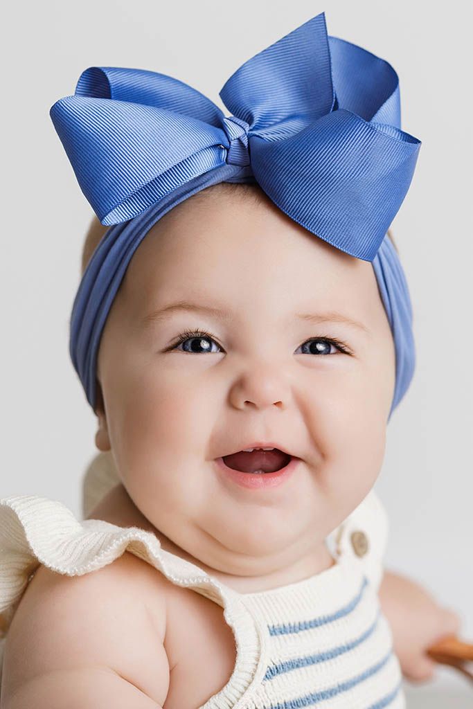 Adorable smiling baby girl wearing large blue bow headband and white striped ruffle outfit.
