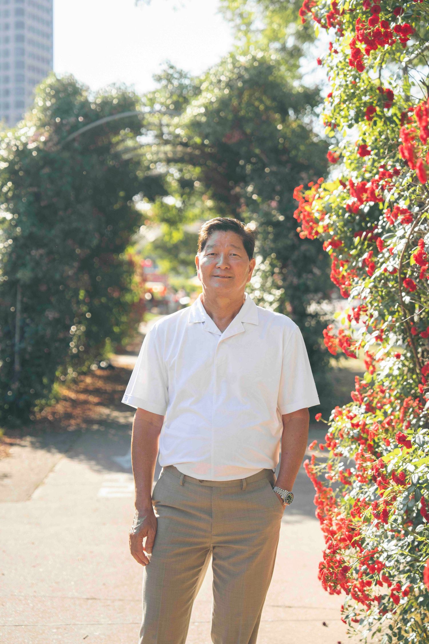 Middle-aged Asian man in white shirt standing in sunny park with red flowering arch.