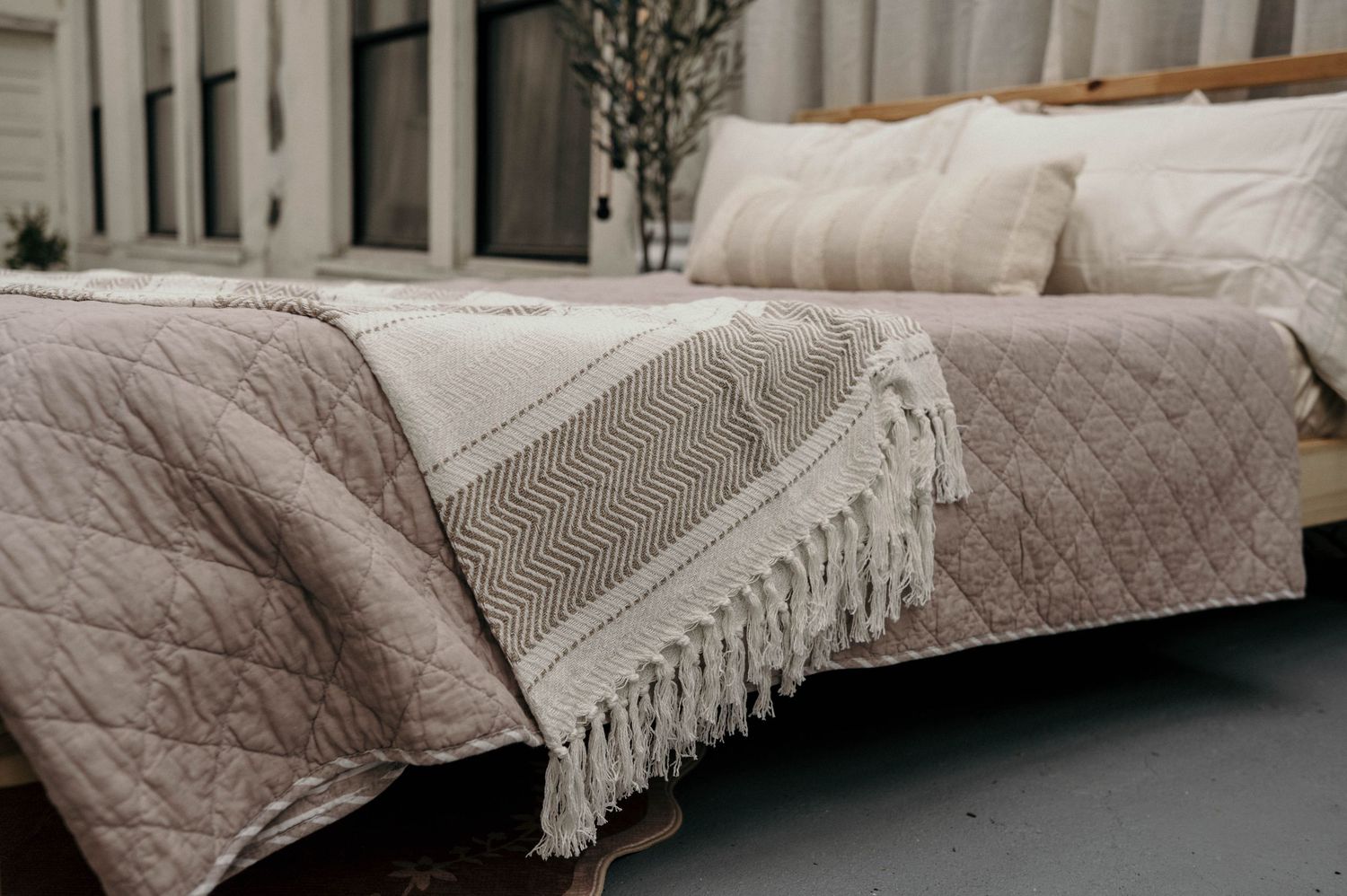 Cozy bed with beige quilted bedding and a fringed woven throw blanket in a neutral-toned bedroom setting.
