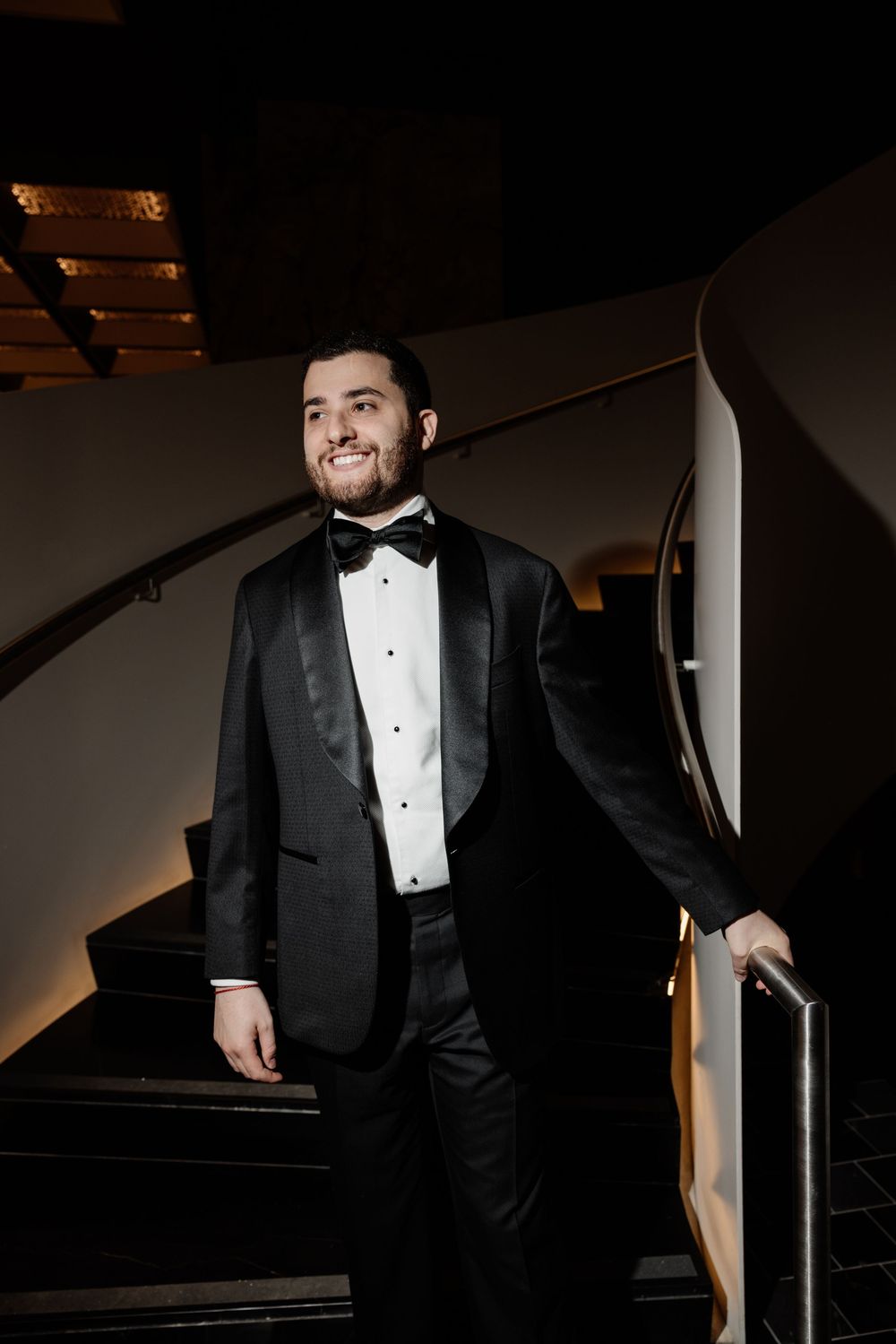 A smiling man in a black tuxedo with bow tie poses on a staircase in an elegant dimly lit venue.