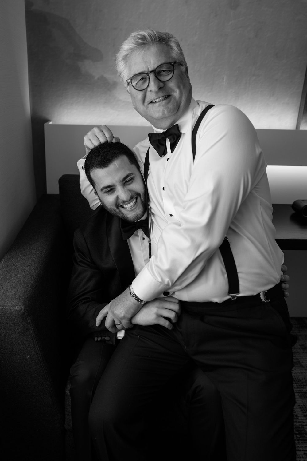 Black and white photo of two men in formal attire smiling together, one older with glasses and suspenders, one younger.