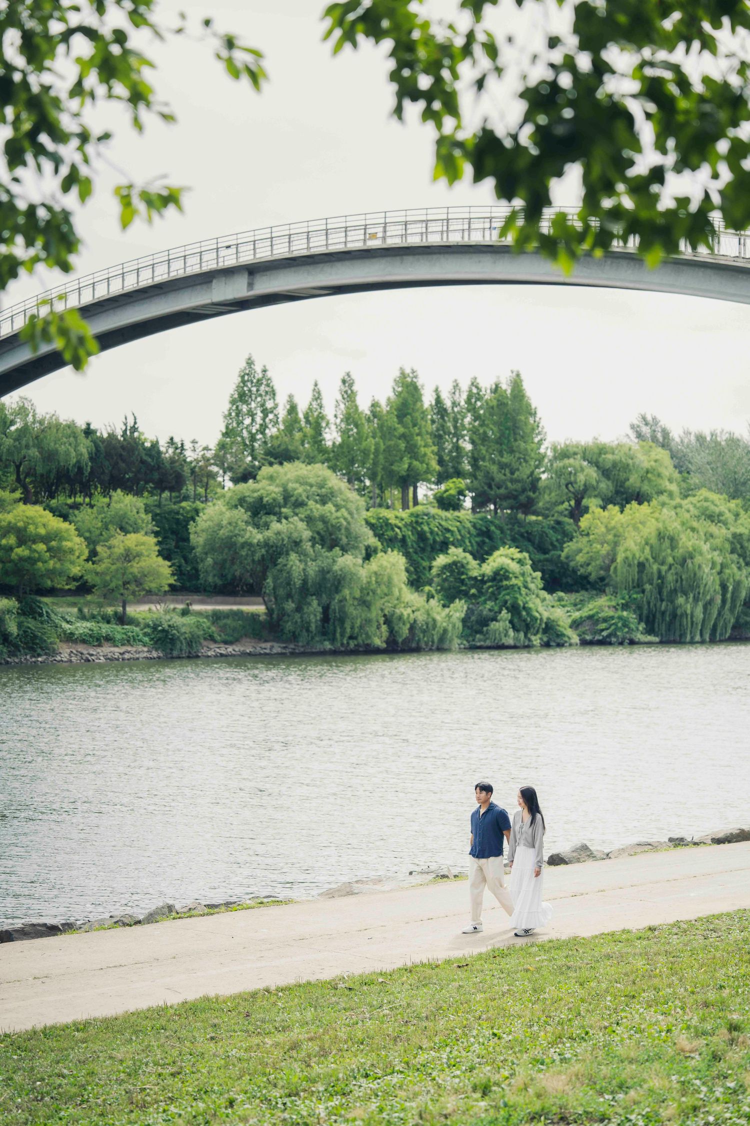 Couple walking hand in hand along a riverside path with an arched bridge and lush green trees in the background.