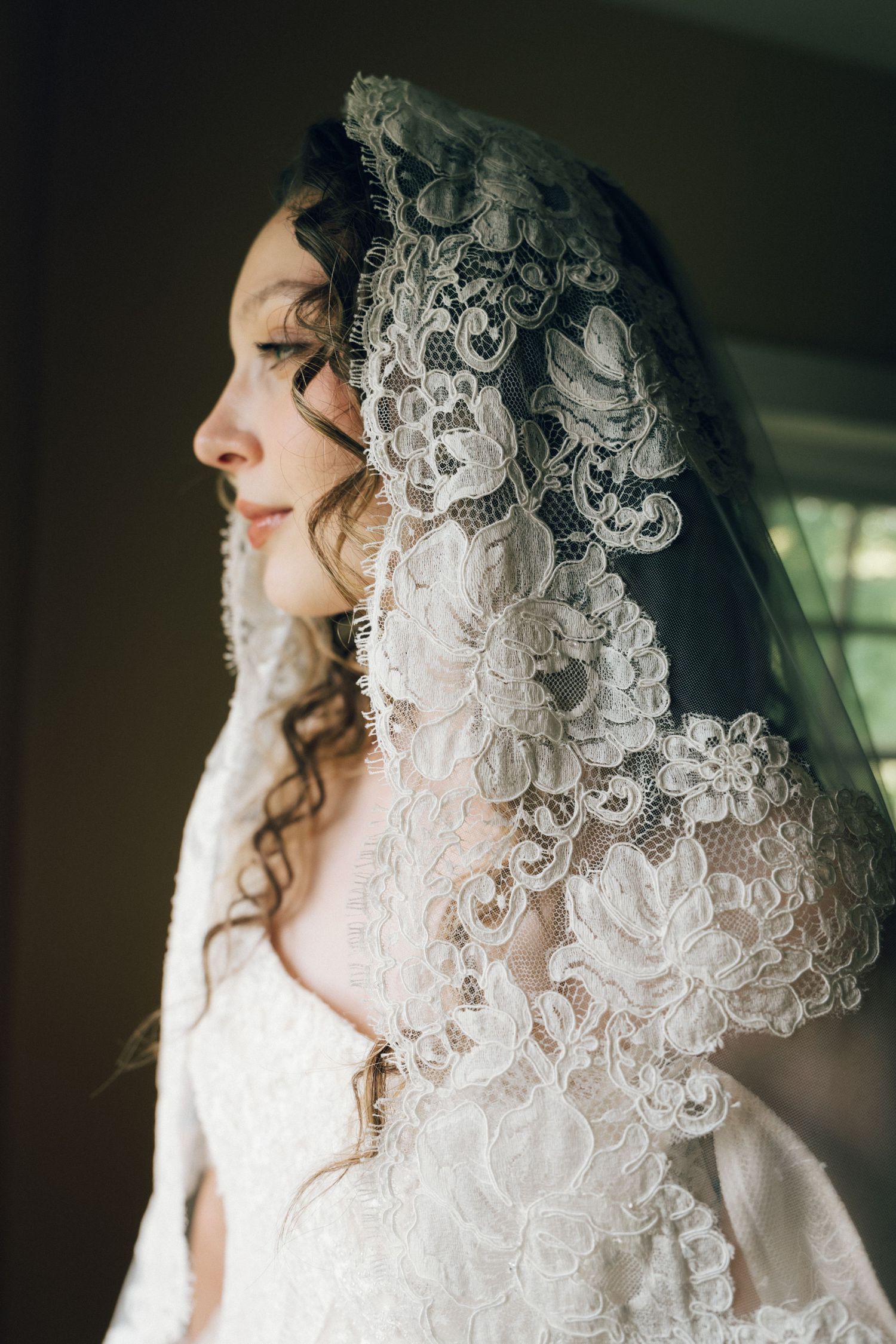 A bride in a white lace wedding dress and intricate floral lace veil stands in soft natural light, gazing serenely.