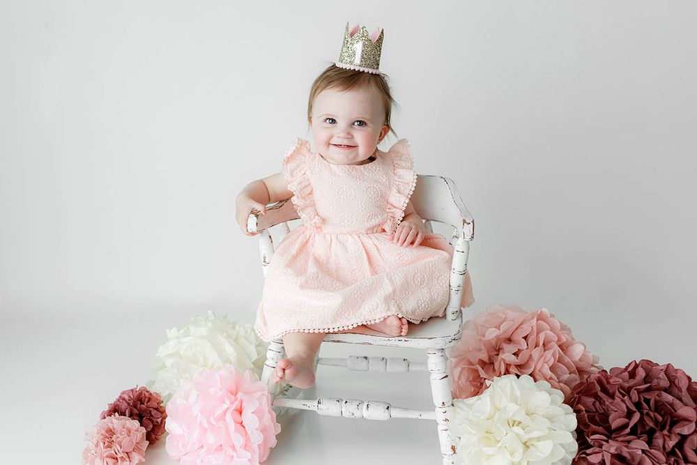 Smiling baby in pink ruffle dress and gold crown sits on white chair with decorative flowers.