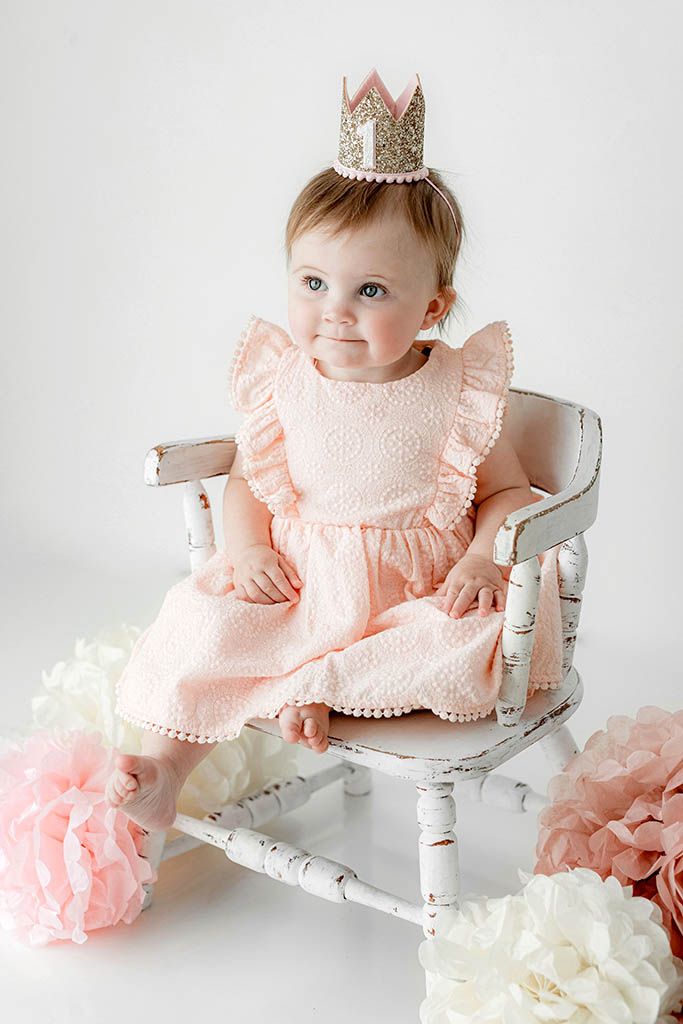 A baby girl in a pink dress and glitter crown sits on a rustic white chair surrounded by pink and white pom poms.