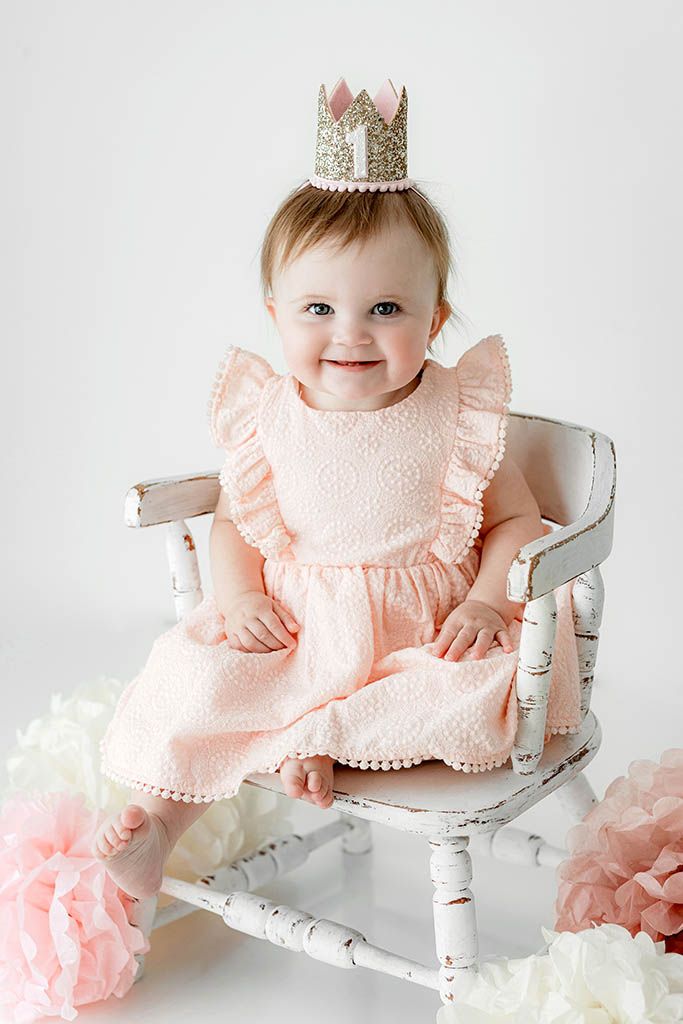 Smiling baby girl in pink dress and gold crown sitting on vintage white chair with floral decorations for first birthday.