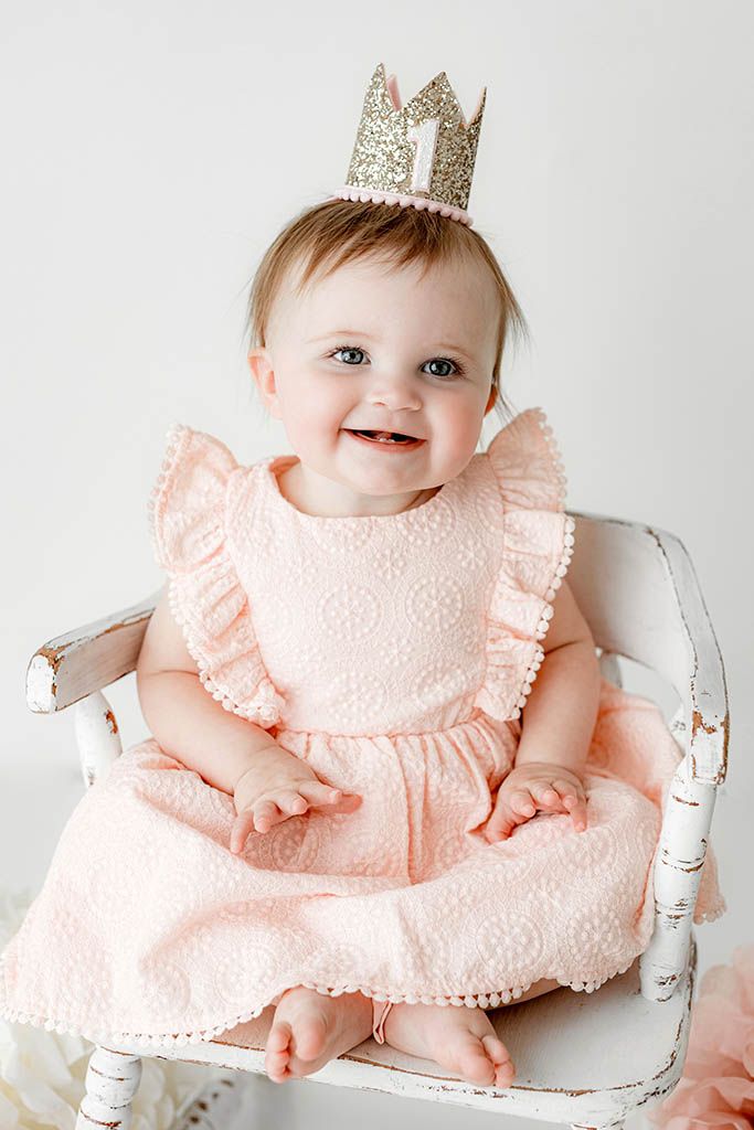 Smiling baby girl in pink lace dress and gold glitter crown sitting in white chair for first birthday photo.