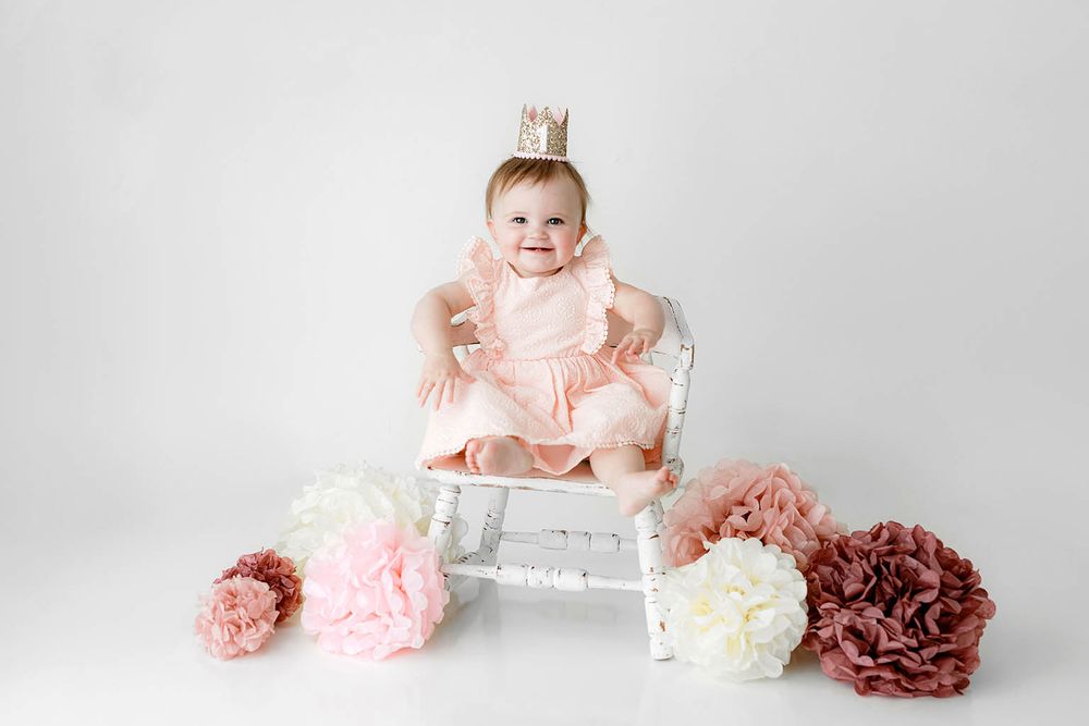 Adorable baby in pink dress and crown laughing on white chair with pink and mauve paper flowers.